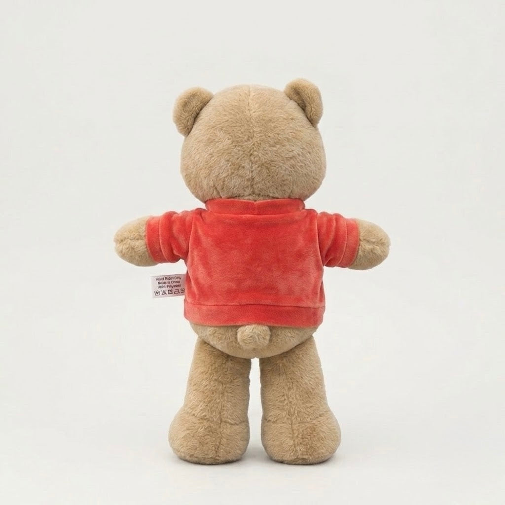 A plush teddy bear viewed from the back, wearing a bright red long-sleeve shirt. The bear is made of soft tan plush fabric, and a small sewn-in tag is visible on the left side of the red shirt.