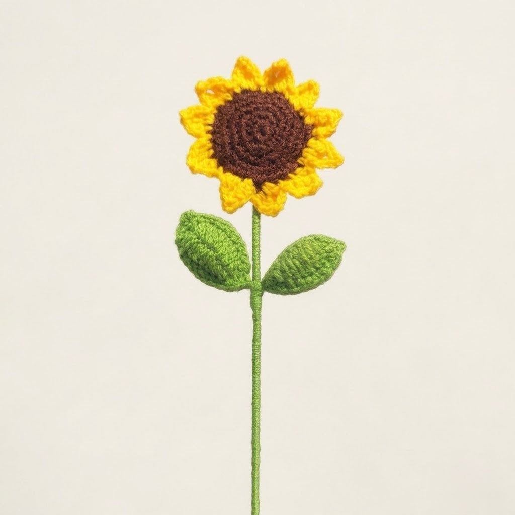 A crocheted sunflower with a yellow and brown flower head, green stem, and two green leaves, centered against a plain light beige background.