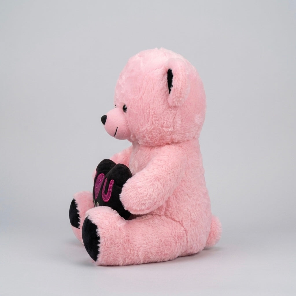 A pink plush teddy bear sitting down, holding a black heart with pink embroidered letters, set against a plain light gray background.