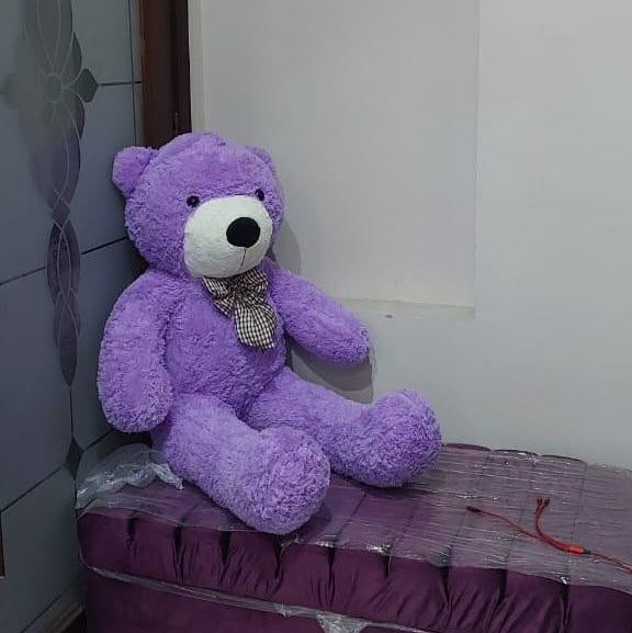Purple teddy bear with a checkered bow tie sitting on a bed in a room.