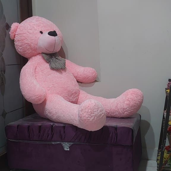 Large pink teddy bear sitting on a purple ottoman against a white wall.