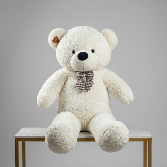 A large plush white teddy bear sitting on a white marble-top table with gold legs, set against a solid gray studio background. The bear has a small brown heart-shaped tag on its left ear and wears a brown and white checkered bow tie.