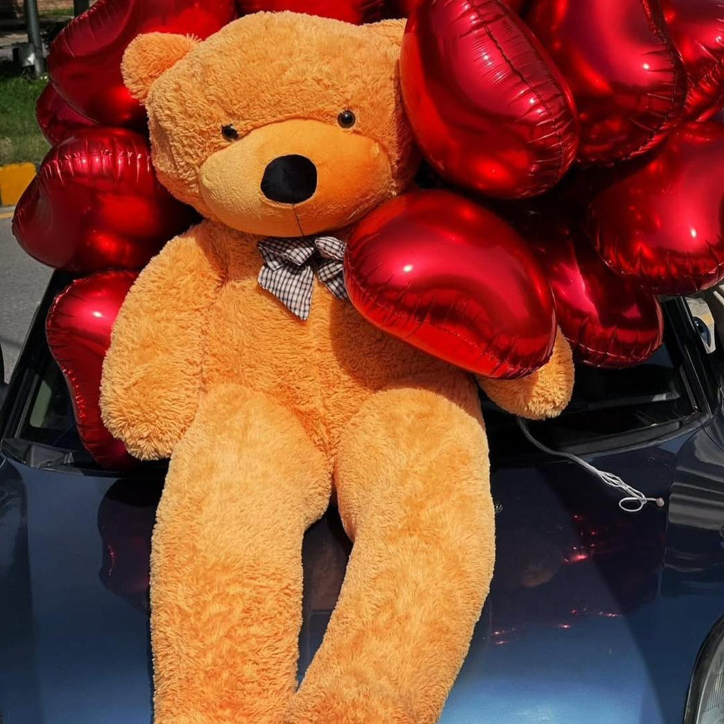 Teddy bear with a bow tie surrounded by red heart-shaped balloons on a car hood.