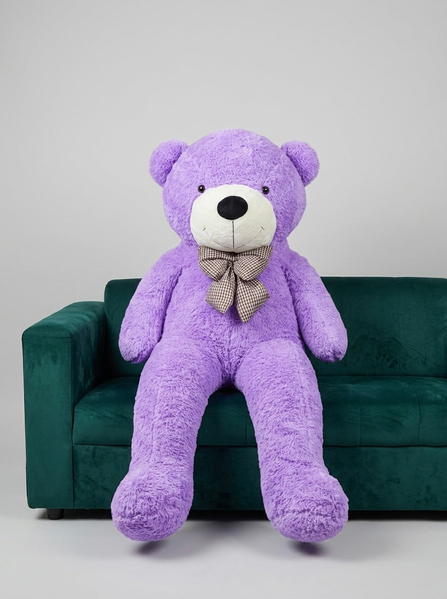 A large purple plush teddy bear with a white snout and black nose, wearing a gray patterned bow tie, sitting on a dark green velvet sofa against a light gray studio background.