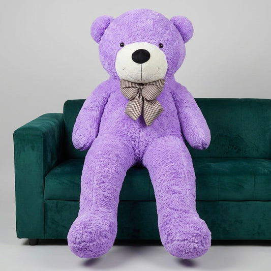 A large purple plush teddy bear with a white snout and black nose, wearing a gray patterned bow tie, sitting on a dark green velvet sofa against a light gray studio background.