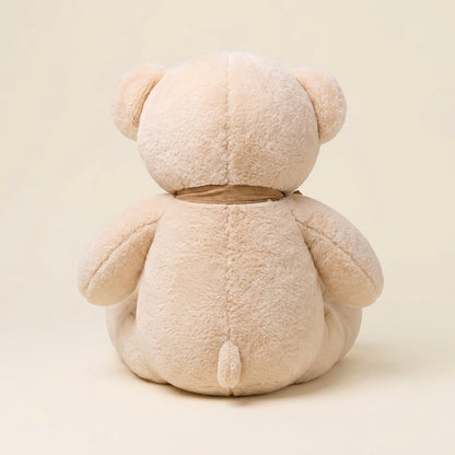 A plush teddy bear toy shown from the back, made of soft beige plush fabric with a small brown ribbon tied around its neck, set against a solid cream background.