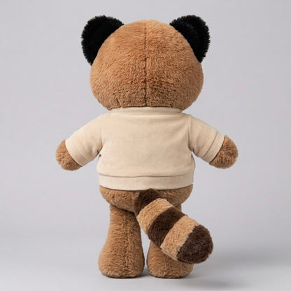 A plush toy animal shown from the back, featuring a brown furry body, black ear patches, a cream-colored short-sleeve shirt, and a brown-and-cream striped tail, set against a plain light gray studio background.