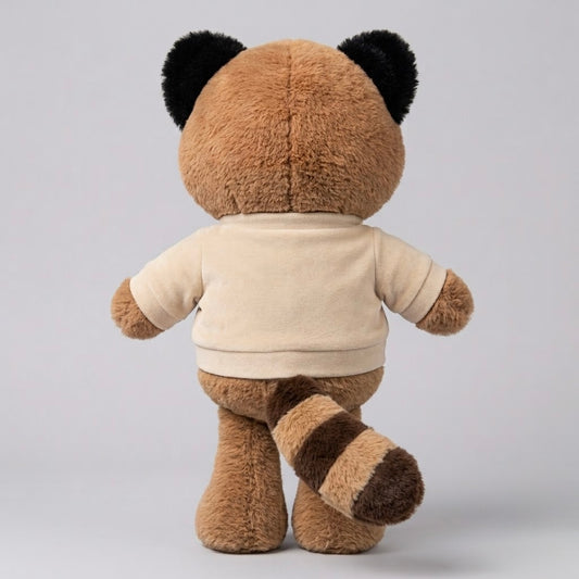 A plush toy animal shown from the back, featuring a brown furry body, black ear patches, a cream-colored short-sleeve shirt, and a brown-and-cream striped tail, set against a plain light gray studio background.