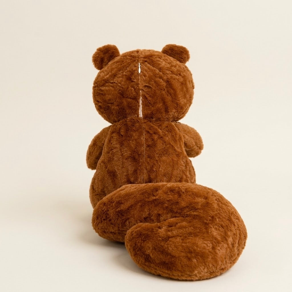 A brown plush teddy bear sitting with its back to the camera, showing a soft textured fur surface and a vertical seam down the center, set against a plain off-white background.