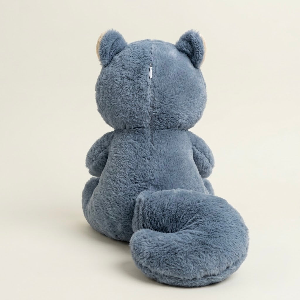 A plush blue-gray bear toy shown from the back, featuring soft textured fur, small upright ears, and a long curled tail, photographed against a plain light beige studio background.