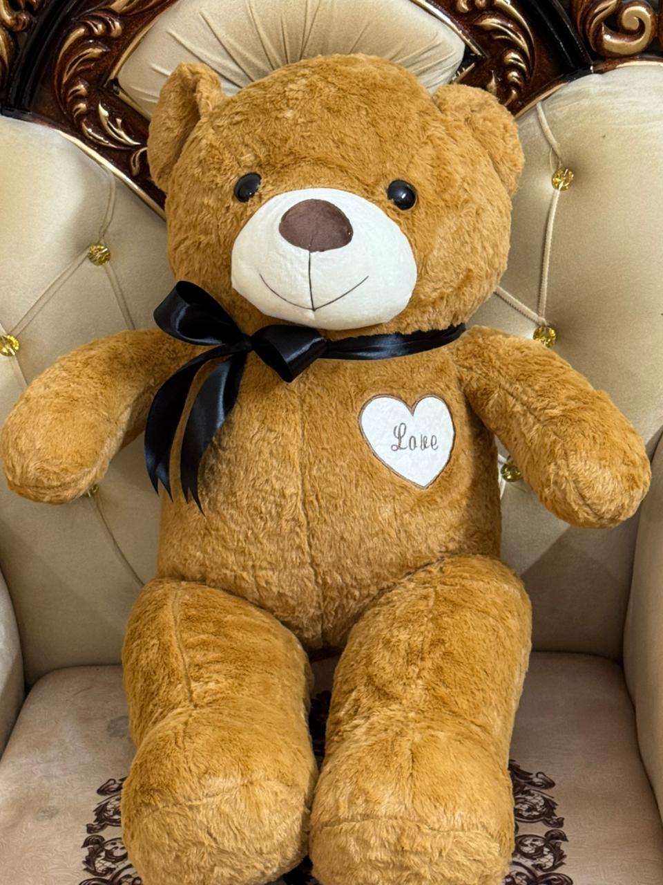 Brown teddy bear with a black bow and 'Love' heart on a decorative chair.