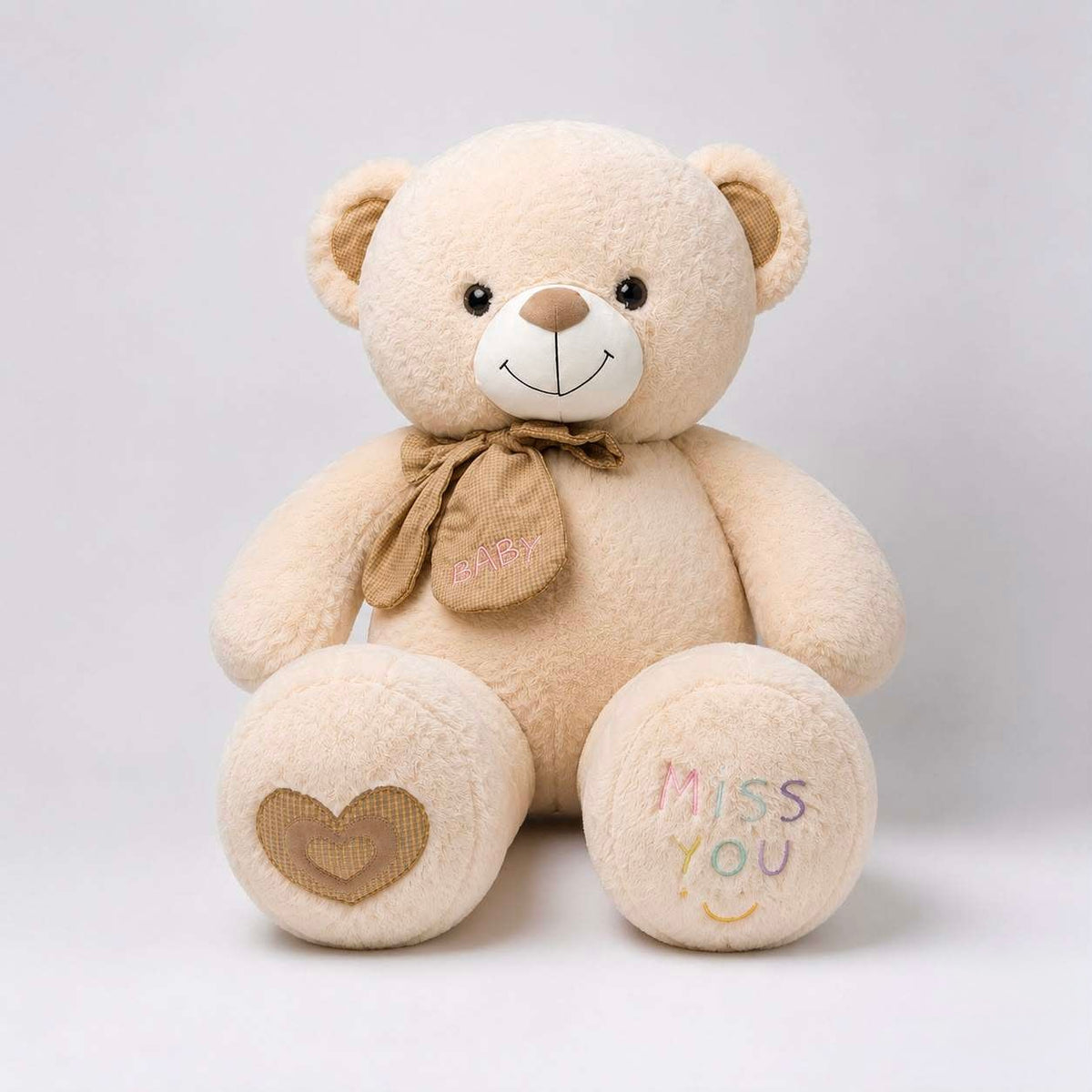 A beige plush teddy bear sitting against a light gray background. The bear has embroidered details, including a brown bow with 'HAPPY' on it, a brown heart on one paw, and 'MISS YOU' with a smiley face on the other paw.