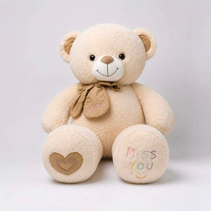 A beige plush teddy bear sitting against a light gray background. The bear has embroidered details, including a brown bow with 'HAPPY' on it, a brown heart on one paw, and 'MISS YOU' with a smiley face on the other paw.