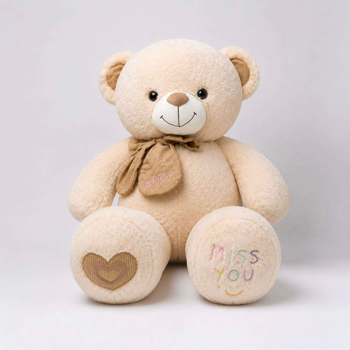 A beige plush teddy bear sitting against a light gray background. The bear has embroidered details, including a brown bow with 'HAPPY' on it, a brown heart on one paw, and 'MISS YOU' with a smiley face on the other paw.