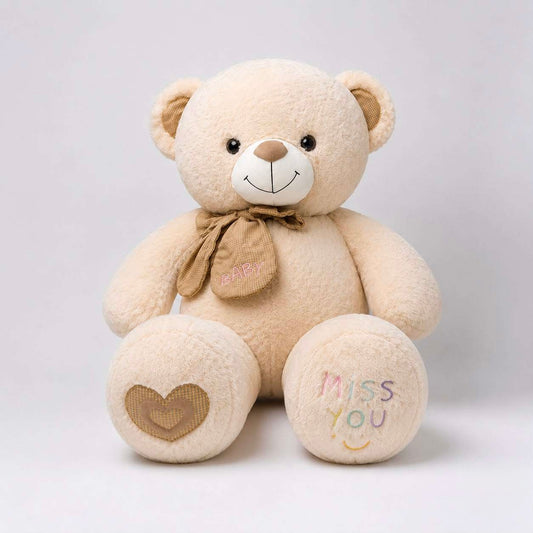 A beige plush teddy bear sitting against a light gray background. The bear has embroidered details, including a brown bow with 'HAPPY' on it, a brown heart on one paw, and 'MISS YOU' with a smiley face on the other paw.
