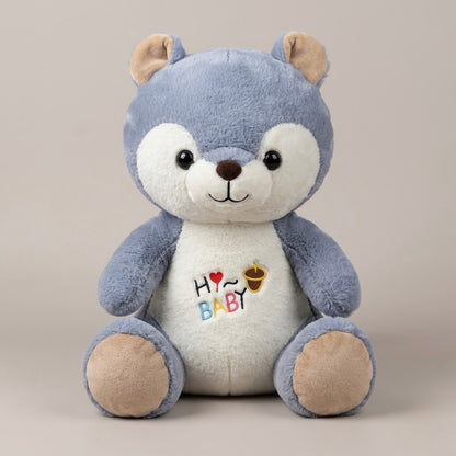 A plush bear toy with a blue-gray body, cream-colored snout and belly, and tan ears and paws. The belly is embroidered with 'HY BABY' in colorful letters and a small acorn graphic.