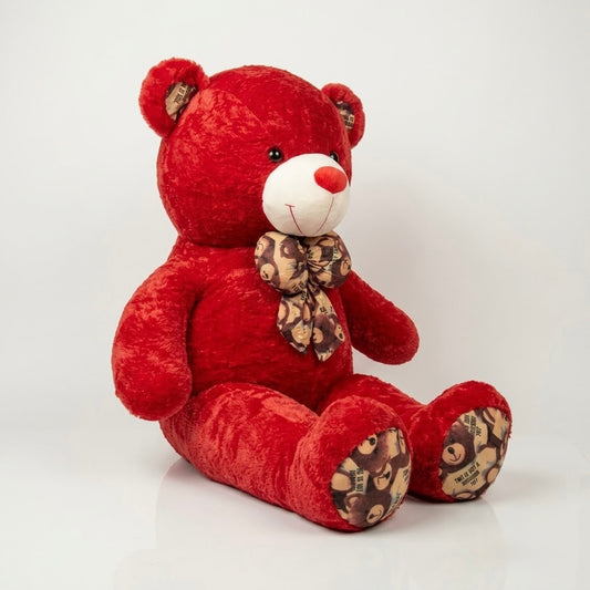 A red plush teddy bear sitting on a white surface against a light gray background. The bear has a white muzzle, black eyes, a red nose, and a brown patterned bow tie.