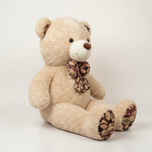 A large beige plush teddy bear sitting on a white studio background, with a brown and beige patterned bow around its neck and patterned fabric on its feet.