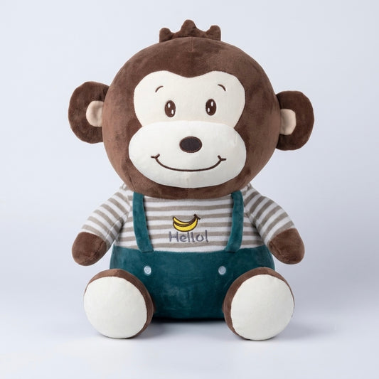 A plush monkey toy sitting upright, with brown fur, a cream-colored face, a striped shirt, and teal overalls. The shirt features a small yellow banana graphic and the word 'Hello!'.