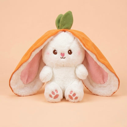 A white plush bunny toy styled like a carrot, with long floppy orange outer ears, pink inner ears, a green leaf on top, and pink paw pads, centered on a soft peach background.