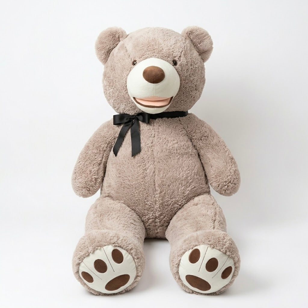 A large plush teddy bear sitting upright against a plain light gray studio background. The bear is taupe-brown with a cream-colored snout, paw pads, and ear areas, a dark brown nose, and a black ribbon bow around its neck.