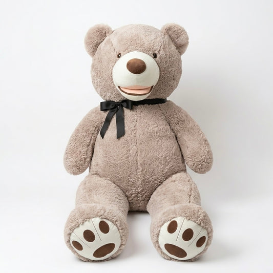 A large plush teddy bear sitting upright against a plain light gray studio background. The bear is taupe-brown with a cream-colored snout, paw pads, and ear areas, a dark brown nose, and a black ribbon bow around its neck.
