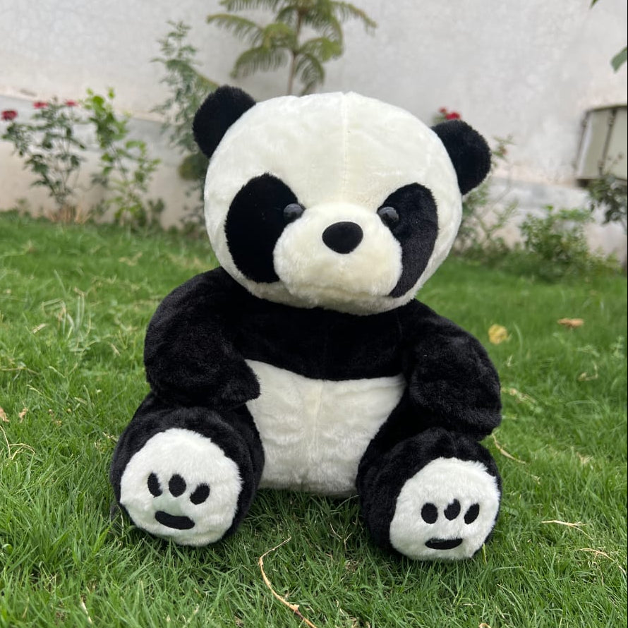 Plush panda toy on grass with a white wall and plants in the background