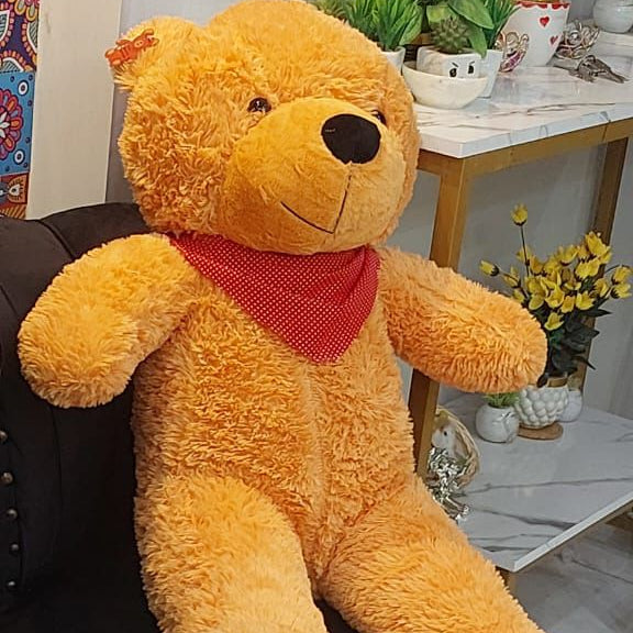 Large teddy bear with a red scarf sitting on a black chair in a room with plants and furniture.