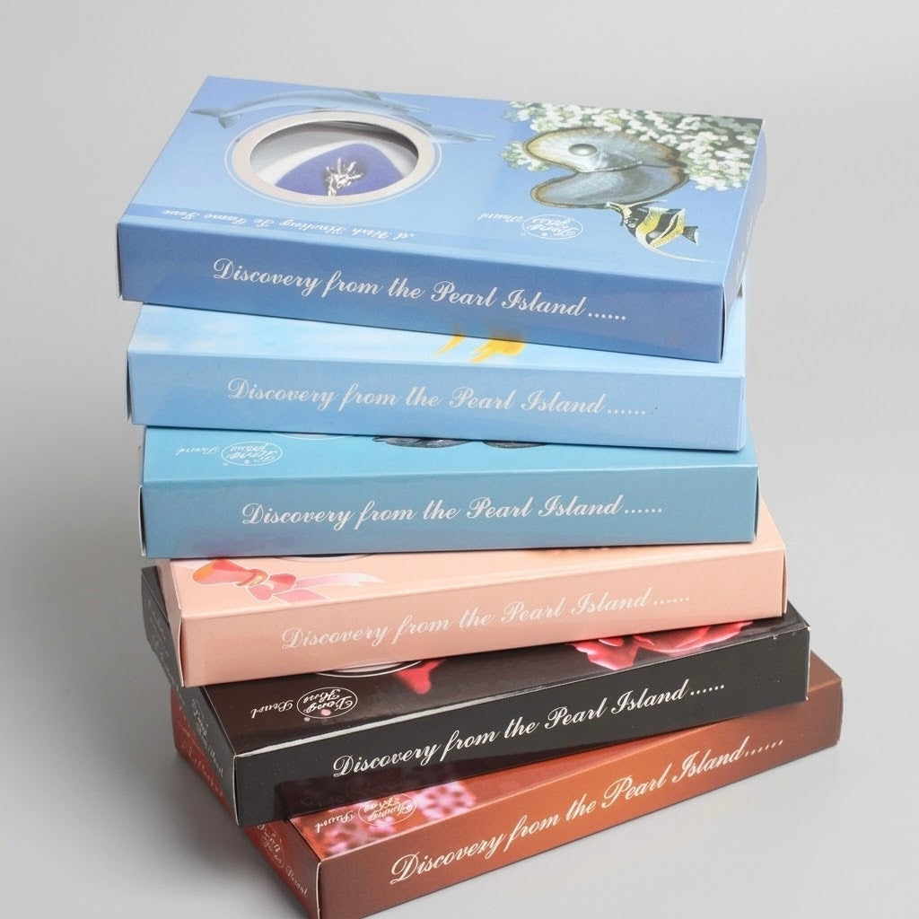 A stack of five decorative product boxes on a light gray background. The boxes are in different colors: light blue, medium blue, pale pink, black, and dark brown, each printed with the phrase 'Discovery from the Pearl Island.