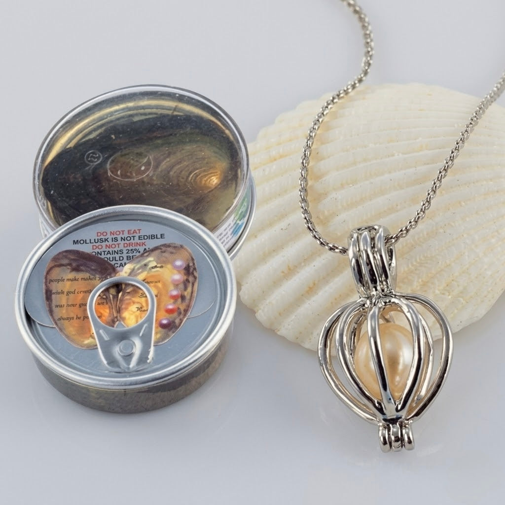 A silver-toned necklace with a heart-shaped pendant containing a pearl-like bead, displayed next to a small round tin with a pull-tab lid and a white seashell on a light gray background.