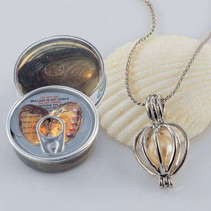 A silver-toned necklace with a heart-shaped pendant containing a pearl-like bead, displayed next to a small round tin with a pull-tab lid and a white seashell on a light gray background.