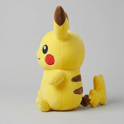A yellow Pikachu plush toy sitting on a light gray studio background, with brown ear and tail accents, a red circular cheek patch, and a small black eye detail.