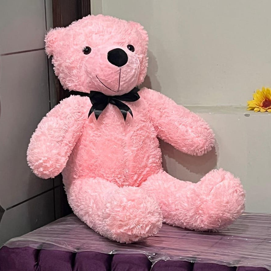 Pink teddy bear with a black bow tie sitting on a glass table against a tiled wall.