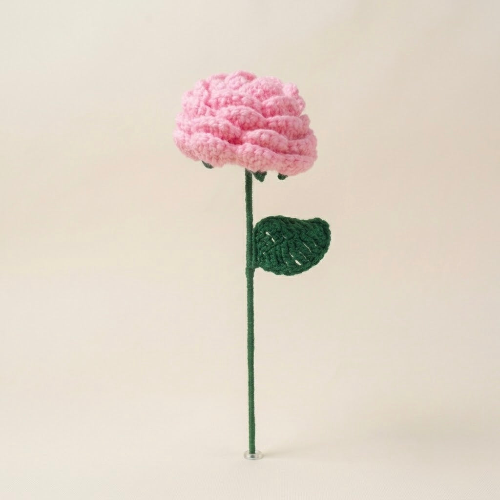 A crocheted flower stem with a pink textured bloom and a green stem and leaf, displayed against a plain light beige background.