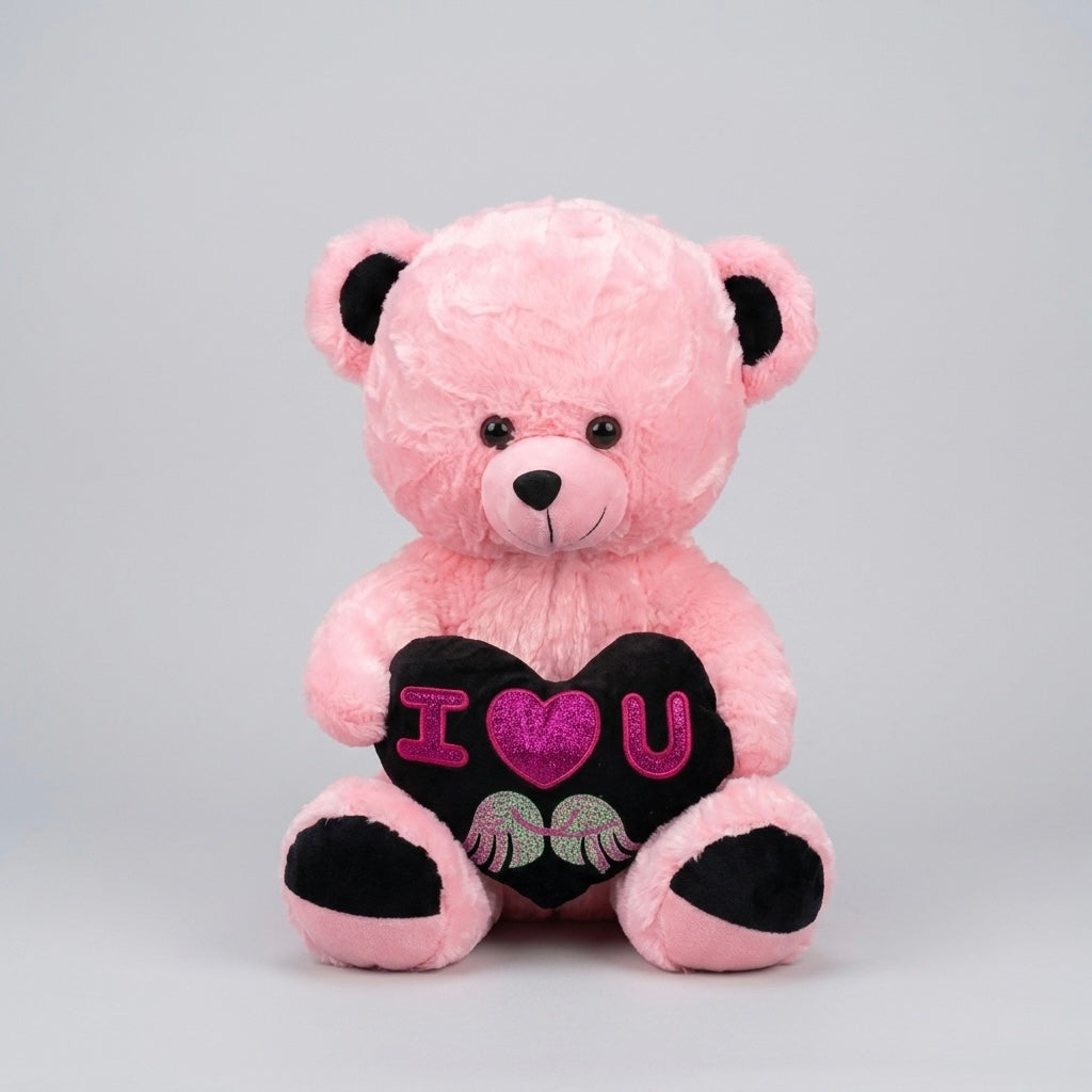 A pink plush teddy bear sitting and holding a black heart-shaped pillow that reads 'I <heart> U' with pink glittery lettering and small embroidered details, photographed against a light gray studio background.