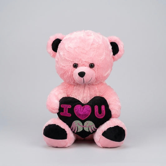 A pink plush teddy bear sitting and holding a black heart-shaped pillow that reads 'I <heart> U' with pink glittery lettering and small embroidered details, photographed against a light gray studio background.