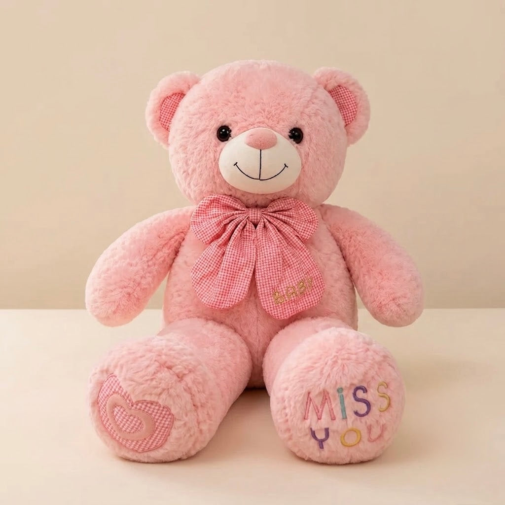 A pink plush teddy bear sitting on a light beige surface against a matching beige background. The bear has a white snout and black eyes, wearing a pink gingham bow with gold embroidered letters.