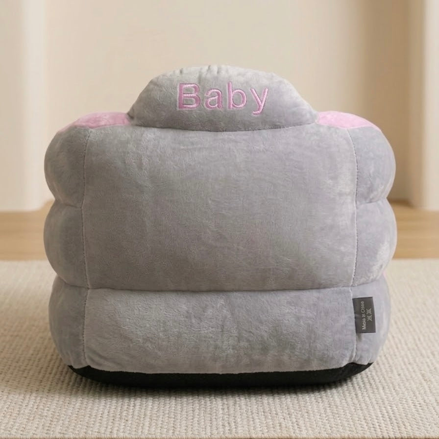A plush gray baby bean bag chair with pink accents, featuring the word 'Baby' embroidered on the top and a small black brand tag on the side, set on a textured beige rug.
