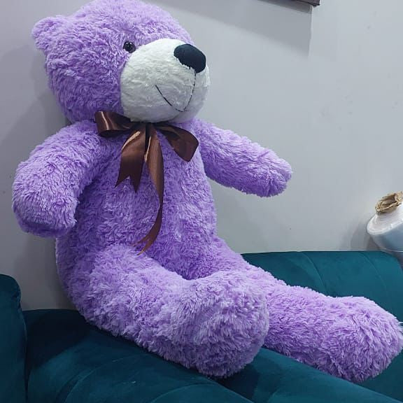 Purple teddy bear with a brown bow on a teal couch, with a framed picture of ducks in the background.