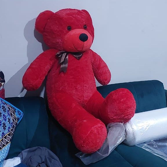 Red teddy bear with a bow tie on a couch with a painting of children and animals in the background.