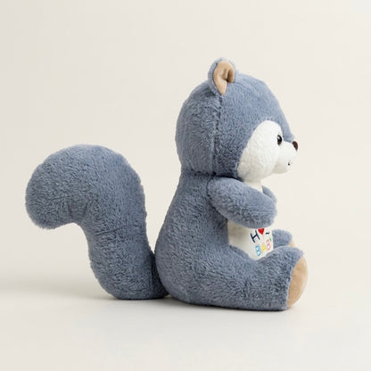 A plush toy squirrel in a dusty blue-gray color with a white face and belly, sitting and holding a small colorful heart-shaped card, photographed against a clean off-white background.