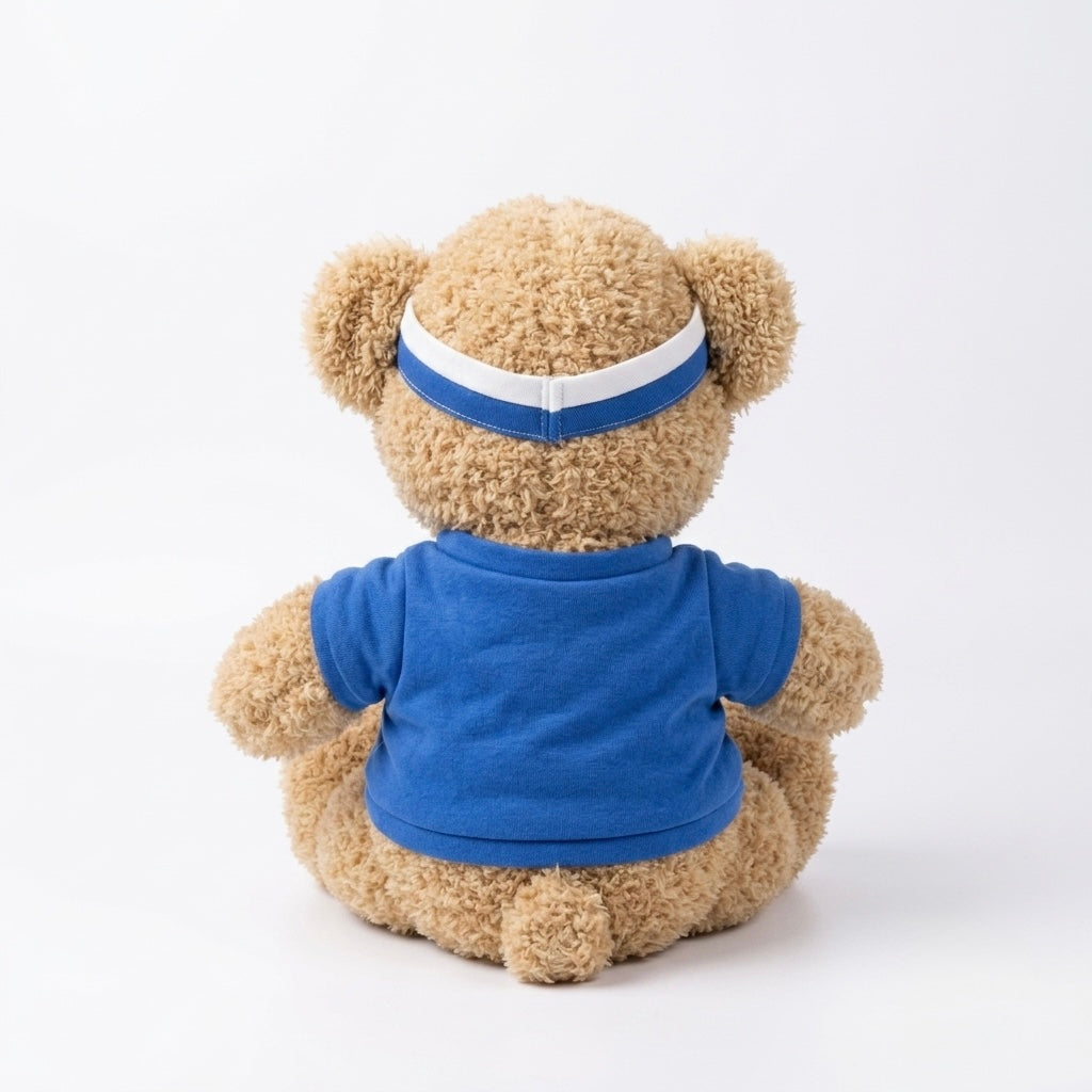 A plush teddy bear viewed from the back, wearing a royal blue short-sleeve shirt and a blue headband with a white stripe, set against a plain white studio background.