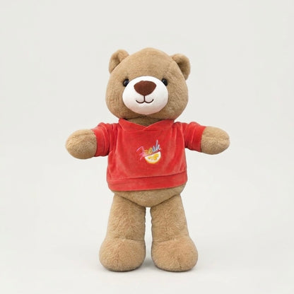 A plush brown teddy bear wearing a red short-sleeve shirt with a small colorful graphic on the chest, standing against a plain light gray background.