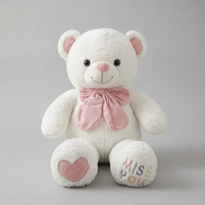 A white plush teddy bear with pink inner ears, a pink bow tie, a pink heart patch on one foot, and 'Miss You' embroidered on the other foot, set against a neutral gray background.