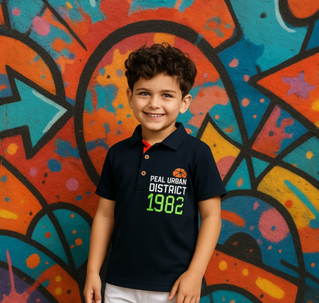 Child wearing a black polo shirt with text standing in front of a colorful graffiti wall.