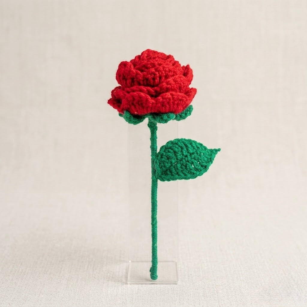 A crocheted red rose with green stem, leaves, and a clear acrylic stand, photographed against a light beige textured background.