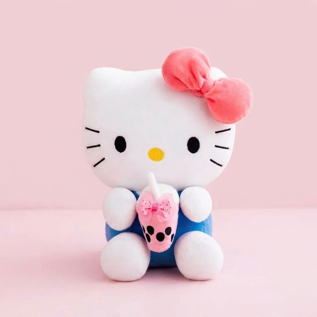 A Hello Kitty plush toy sitting upright, featuring her signature white face and ears, a pink bow, black whiskers, a yellow nose, and blue pants. She is holding a small pink drink cup with a bow on a pink background.